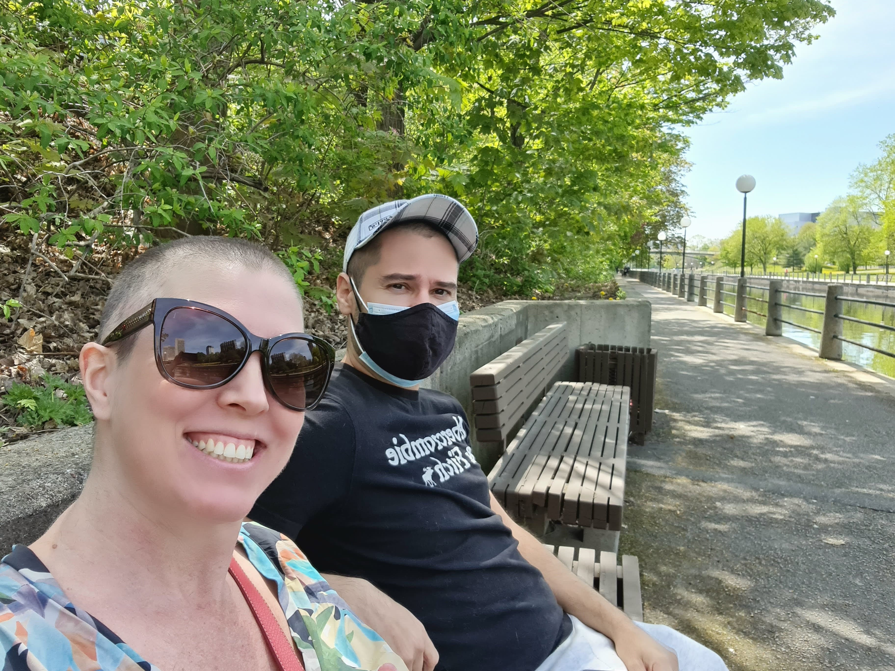A woman and man sit outdoors on a park bench. The woman is smiling at the camera, wearing close-cropped hair and sunglasses. The man is wearing a face mask and a baseball cap.